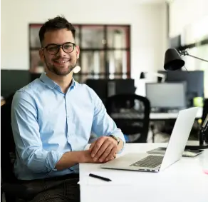 A imagem mostra um homem sorridente, em um escritório, usando seu laptop.