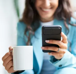 A imagem mostra uma mulher, segurando uma caneca de porcelana e usando o seu celular.