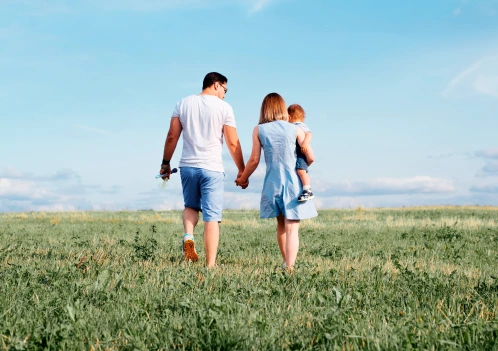 Família caminhando de mãos dadas em um campo verde, com uma criança nos braços da mãe e céu azul ao fundo.