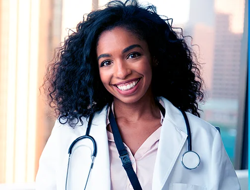 Mulher de cabelo ondulado preto sorrindo, de camisa rosa e jaleco branco e, com um estetoscópio em volta do pescoço.