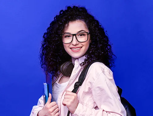 Mulher de cabelo cacheado, óculos e headphone em volta do pescoço, segurando uma pasta na mão direita e mochila na esquerda.