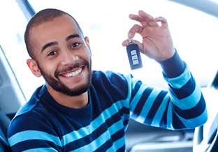 A imagem mostra um rapaz negro, jovem, de barba, no interior de um carro. Ele está do lado do motorista e segura a chave do carro com a mão esquerda enquanto olha pra frente e sorri. Ele está vestindo uma blusa de manga longa azul escura com listras em azul claro.