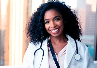 A imagem tem um fundo de prédios desfocados. Em primeiro plano, temos uma mulher jovem, negra, de cabelo ondulado escuro. Ela está sorrindo e veste um jaleco branco e abaixo do jaleco é possível ver uma camisa rosa. Ela está com um estetoscópio em volta do pescoço.