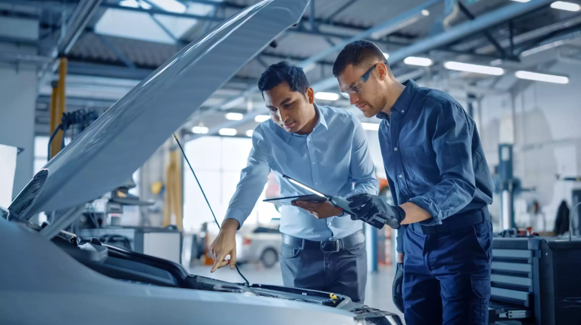 Dois homens inspecionam o motor de um carro em uma oficina mecânica, analisando detalhes.