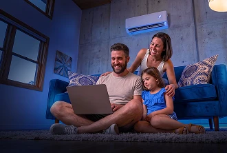 Mulher em sofá azul apoia as mãos nos ombros de um homem e de uma menina, enquanto acompanham juntos algo no computador.