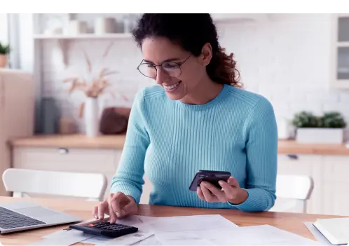 Mulher de suéter azul claro, óculos e cabelos preso, segurando um celular e mexendo em uma calculadora apoiadas em papéis.