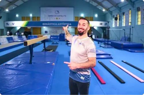 Diego Hypólito em um ginásio de ginástica artística, sorrindo e apontando para os equipamentos do espaço.