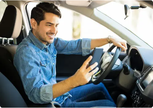 Homem de camisa jeans sorrindo dentro de um carro parado, com uma mão no volante e segurando o celular com a outra.