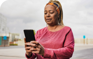 Mulher com faixa xadrez na cabeça e camisa rosa, em um ambiente aberto, segurando e olhando para o celular.