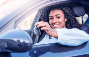 Mulher com cabelo solto e suéter branco, sorrindo dentro de um carro, com o braço para fora segurando uma chave.