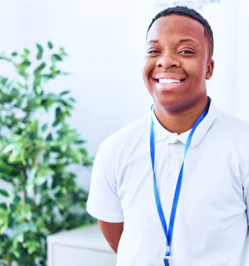 A imagem mostra um homem em um escritório claro. Ele está sorrindo, veste uma camiseta branca e usa um crachá.