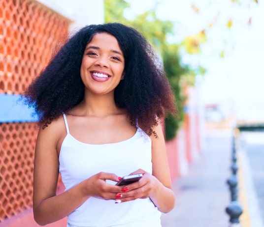 Mulher com cabelo crespo longo, vestindo blusa branca, sorrindo e segurando um celular na mão.