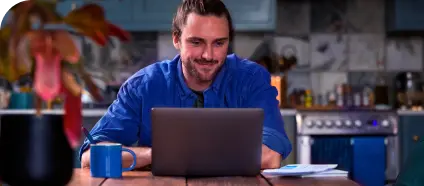 Em uma cozinha, um homem branco de camisa azul esboça um sorriso enquanto olha o computador.