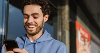 Homem sorridente usando moletom azul e digitando no celular. Ele está na calçada