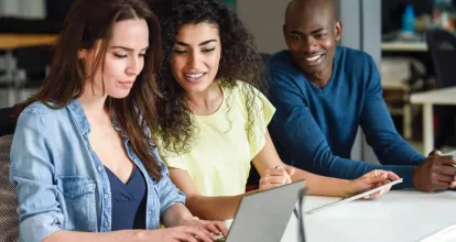 A imagem mostra duas mulheres e um homem sentados. Uma das mulheres está usando um laptop e todos estão olhando.