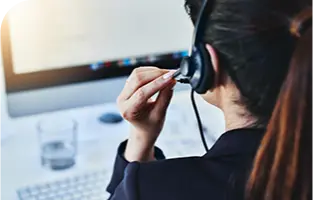 Uma mulher, de costas, falando no microfone do seu headset, em frente à um computador.