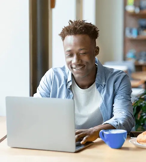 Homem sorrindo, sentado na frente de uma mesa e olhando para a tela de um computador portátil