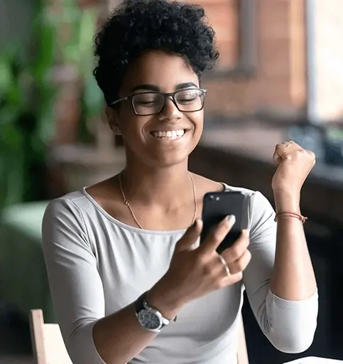 Mulher sorrindo, olhando para a tela do celular com uma das mãos cerradas simbolizando vitória