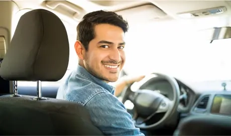 Na imagem há um homem vestido com um jaqueta jeans. Este homem está sorrindo dentro de um carro, que ele está dirigindo.