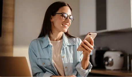 Na imagem há uma mulher de cabelos longos e escuros, blusa social azul claro, sorrindo ao segurar um telefone dourado.