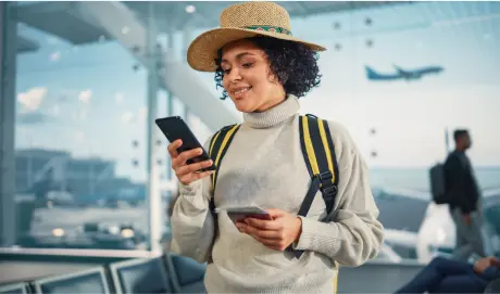 Na imagem há uma mulher em um aeroporto, ela sorri ao mexer no celular.