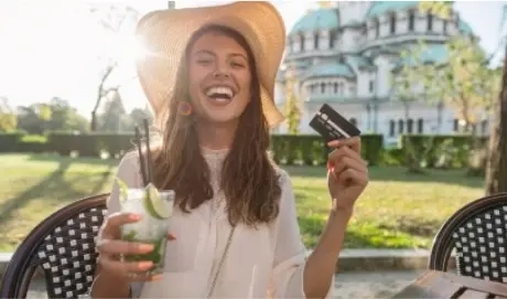 Na imagem há uma mulher de chapéu, ela está sorrindo com uma bebida em uma mão e um cartão na outra.