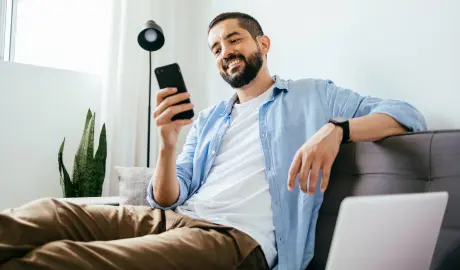 Homem sentado em um sofá cinza, sorrindo ao usar o celular.