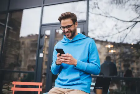 Na imagem há um homem de óculos de grau, ele está em um local aberto, trajado com moletom azul, sorrindo para um celular.