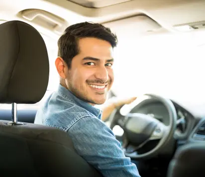 Na imagem há um homem vestido com um jaqueta jeans. Este homem está sorrindo dentro de um carro, que ele está dirigindo.