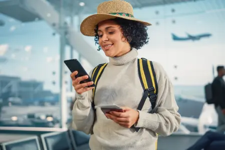 Na imagem há uma mulher em um aeroporto, ela sorri ao mexer no celular.