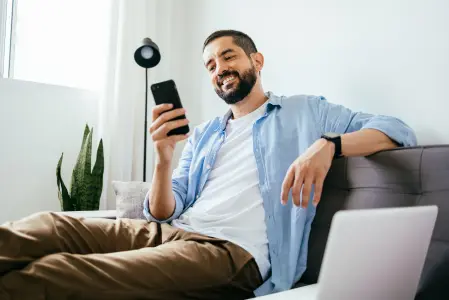 Na imagem há um homem sentado em um sofá mexendo no celular sorrindo.