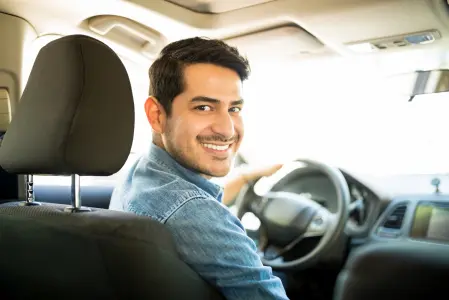 Na imagem há um homem vestido com um jaqueta jeans. Este homem está sorrindo dentro de um carro, que ele está dirigindo.