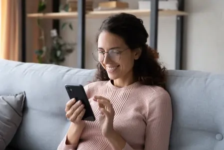 Na imagem há uma mulher sentada em um sofá, ela está mexendo no celular sorrindo.