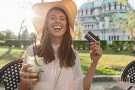 Na imagem há uma mulher de chapéu, ela está sorrindo com uma bebida em uma mão e um cartão na outra.