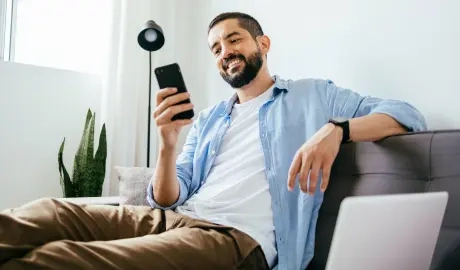 Na imagem há um homem sentado em um sofá mexendo no celular sorrindo.