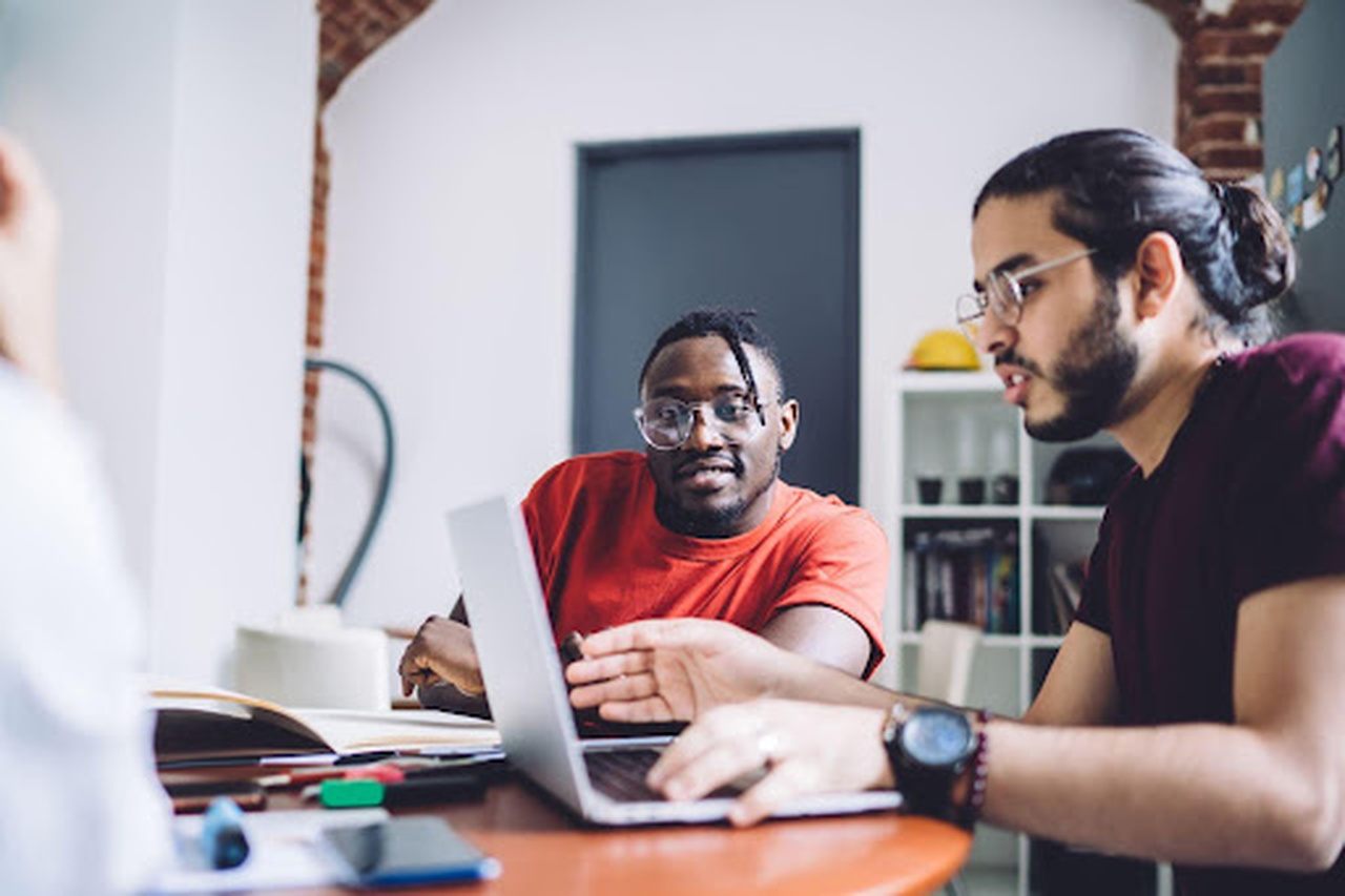 A imagem mostra dois homens sentados a uma mesa e usando um laptop. 