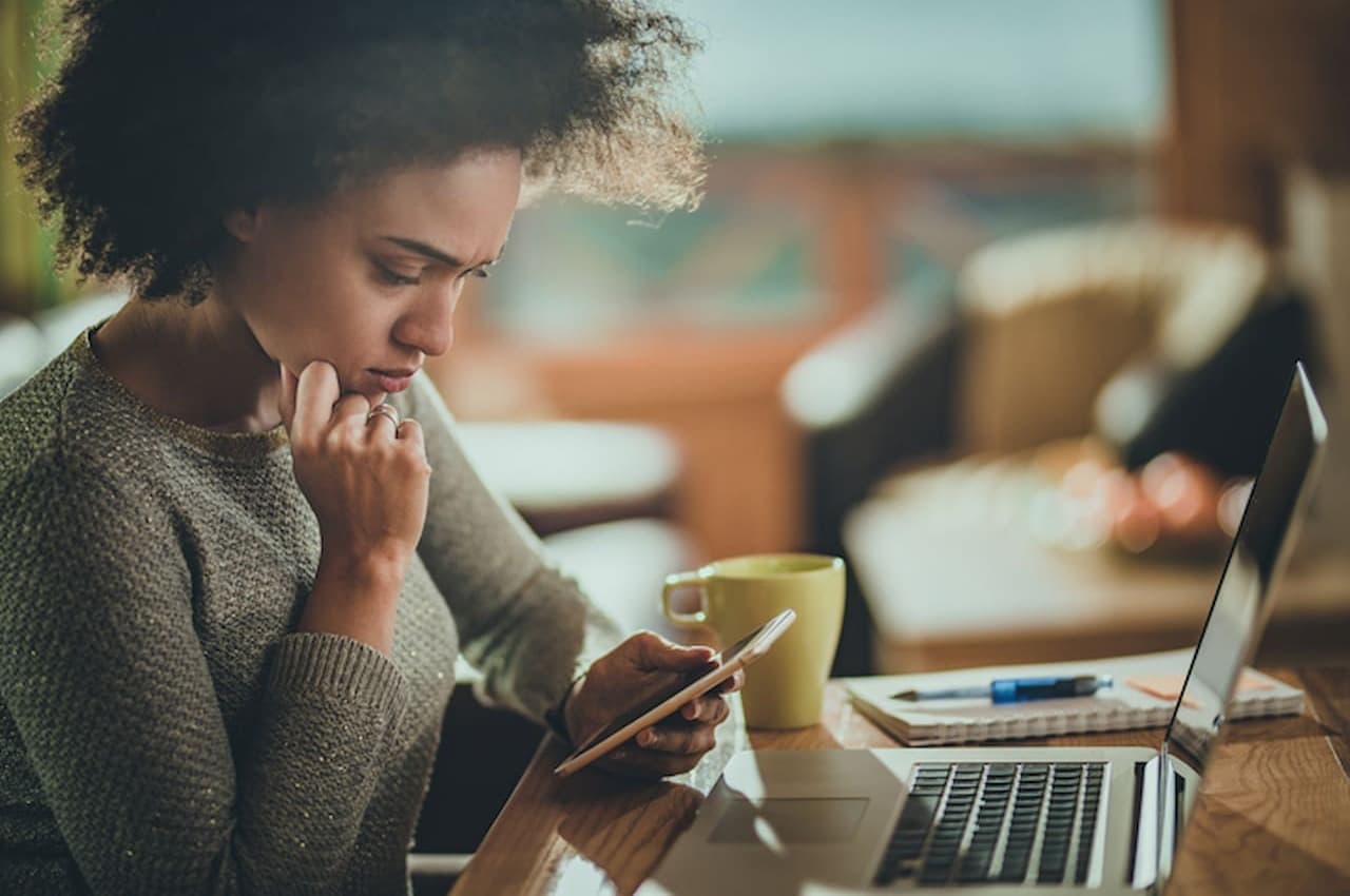 Mulher com expressão concentrada, olhando para o celular e com a mão direita apoiada no rosto. Na mesa a sua frente, tem um computador e blocos de anotação.