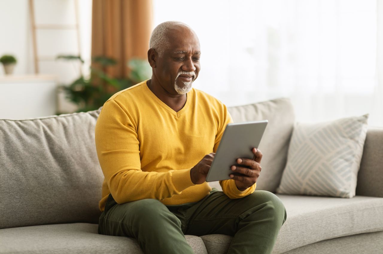 Homem sênior de cabelos curtos grisalhos, utilizando um tablet enquanto está sentado no seu sofá.