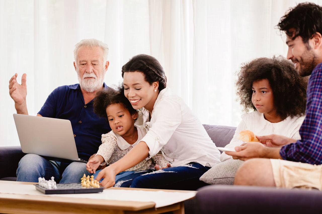 Família composta por um pai, mãe, avós e crianças digitando no celular e com expressões sorridentes. Eles estão no ambiente da sala de estar.