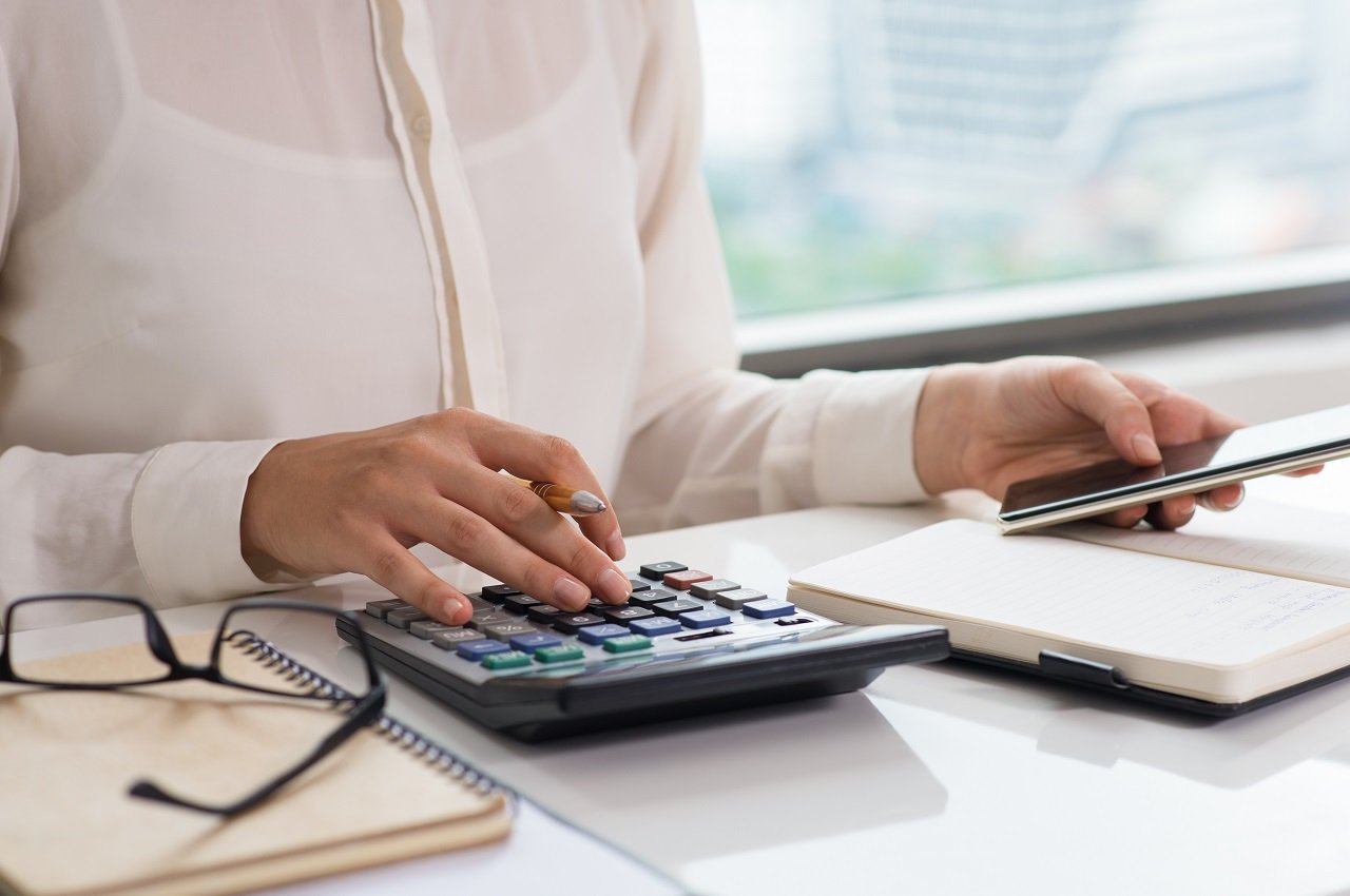 Mulher (seu rosto não aparece na imagem) calculando alíquota na calculadora e utilizando um computador para anotar. Na sua mesa tem caderno de anotações e um óculos de grau.