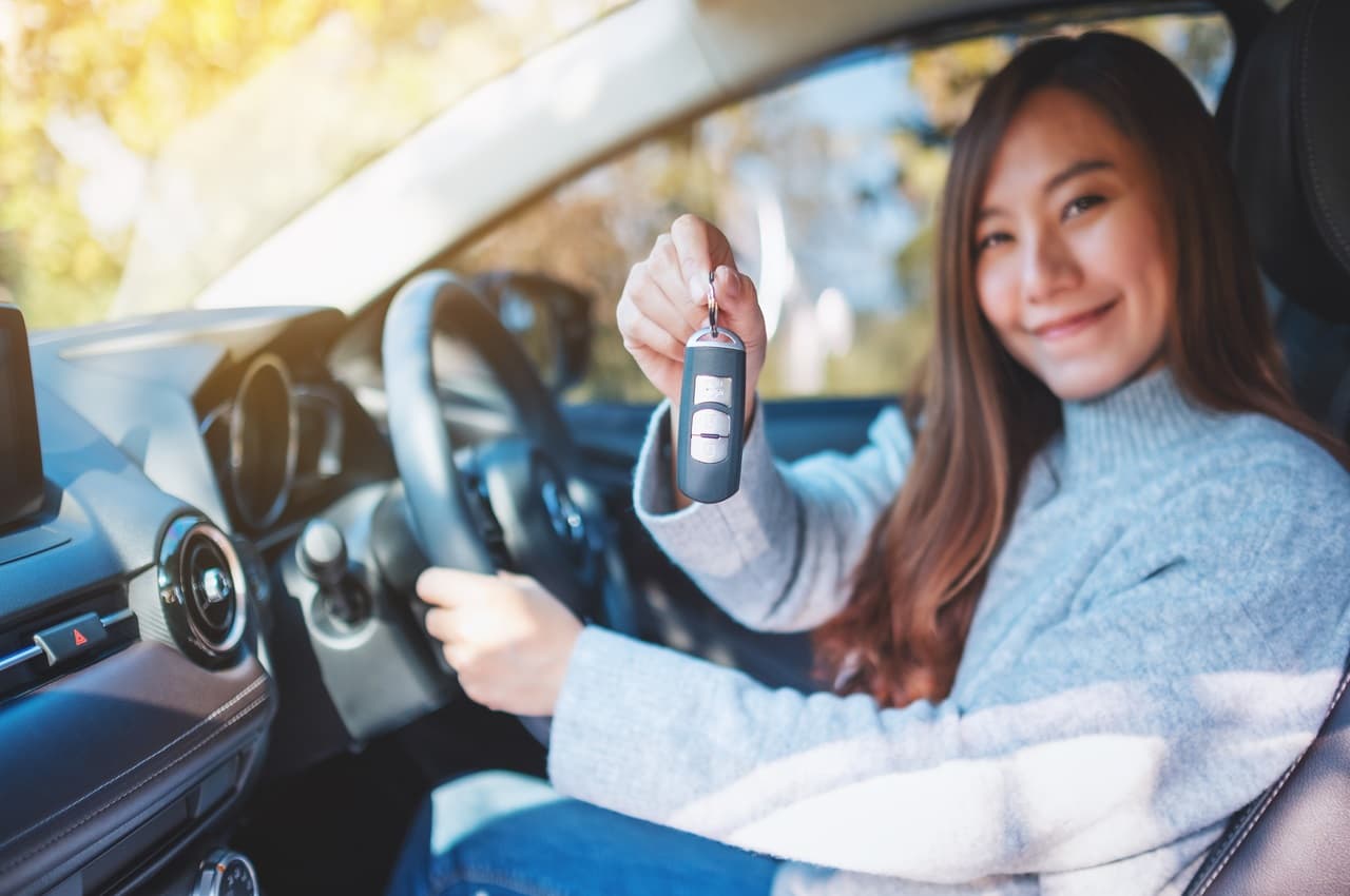 Mulher com expressão sorridente, segurando chaves para alugar ou comprar carro e sentada no banco do motorista.