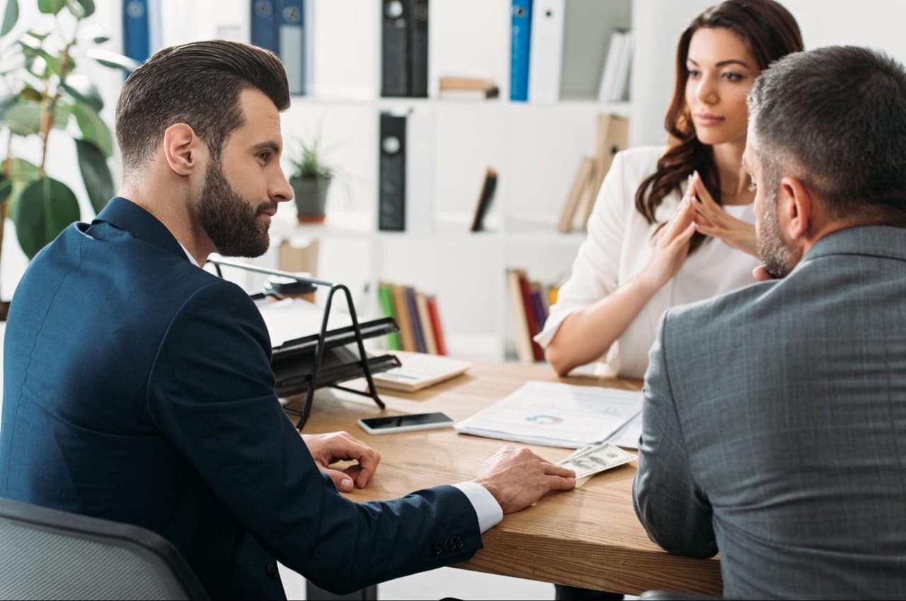 Funcionário solicitando adiantamento salarial para seu empregador. O homem (funcionário) veste terno preto e conversa com uma mulher (chefe).