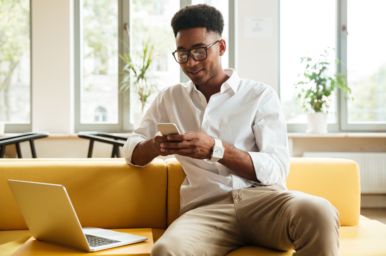 A imagem mostra um homem sentado à um sofá, usando seu celular. Sob o sofá, há um laptop. 