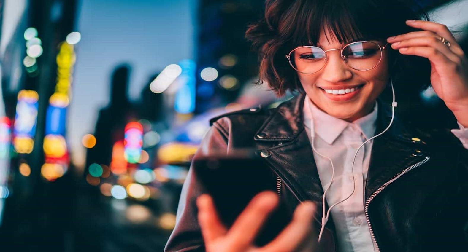 A imagem mostra uma mulher sorridente usando seu celular e seus fones de ouvido.