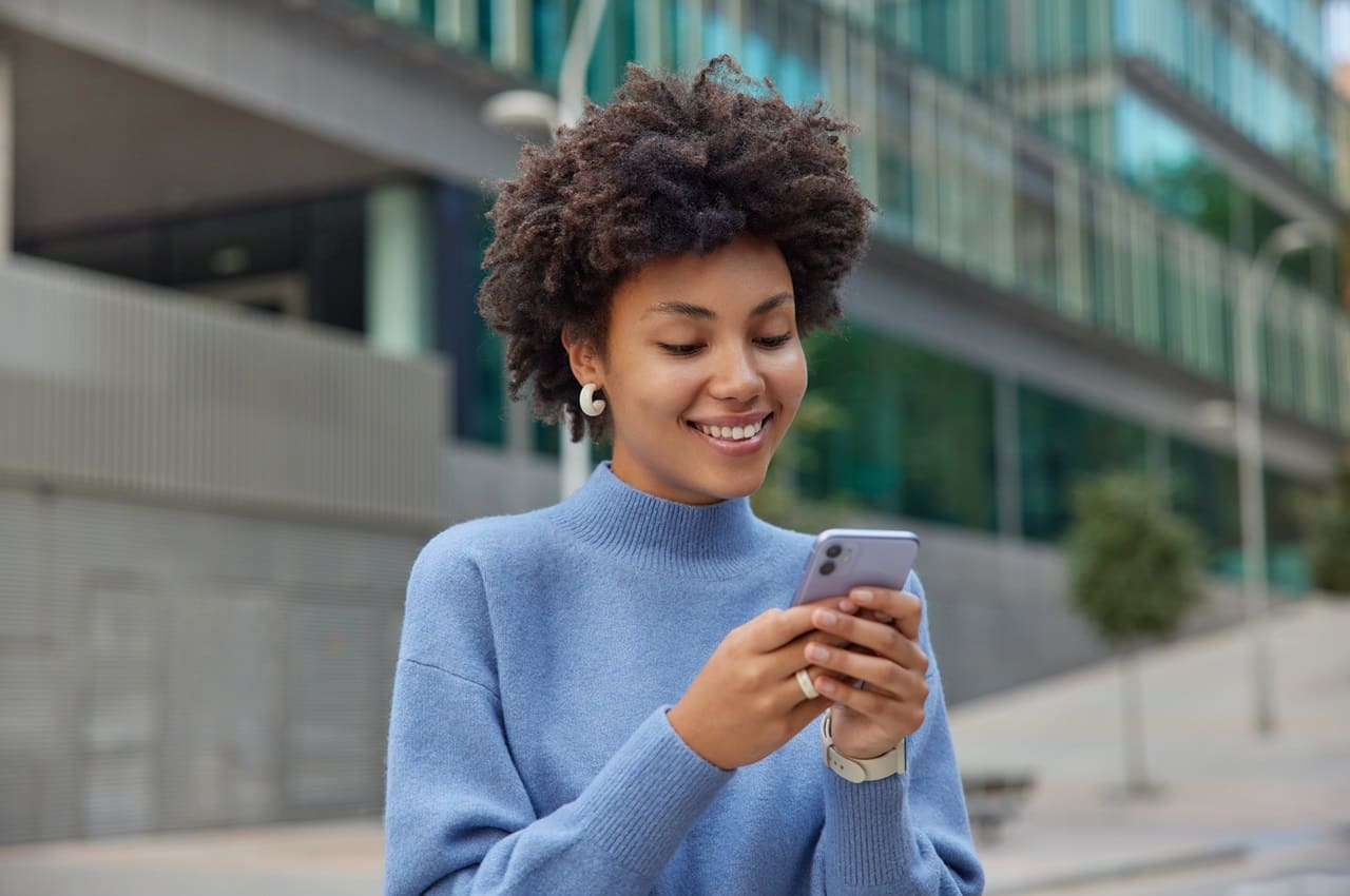 Mulher sorridente em pé na calçada, acessando no celular app darwin seguros. Ela veste suéter azul.