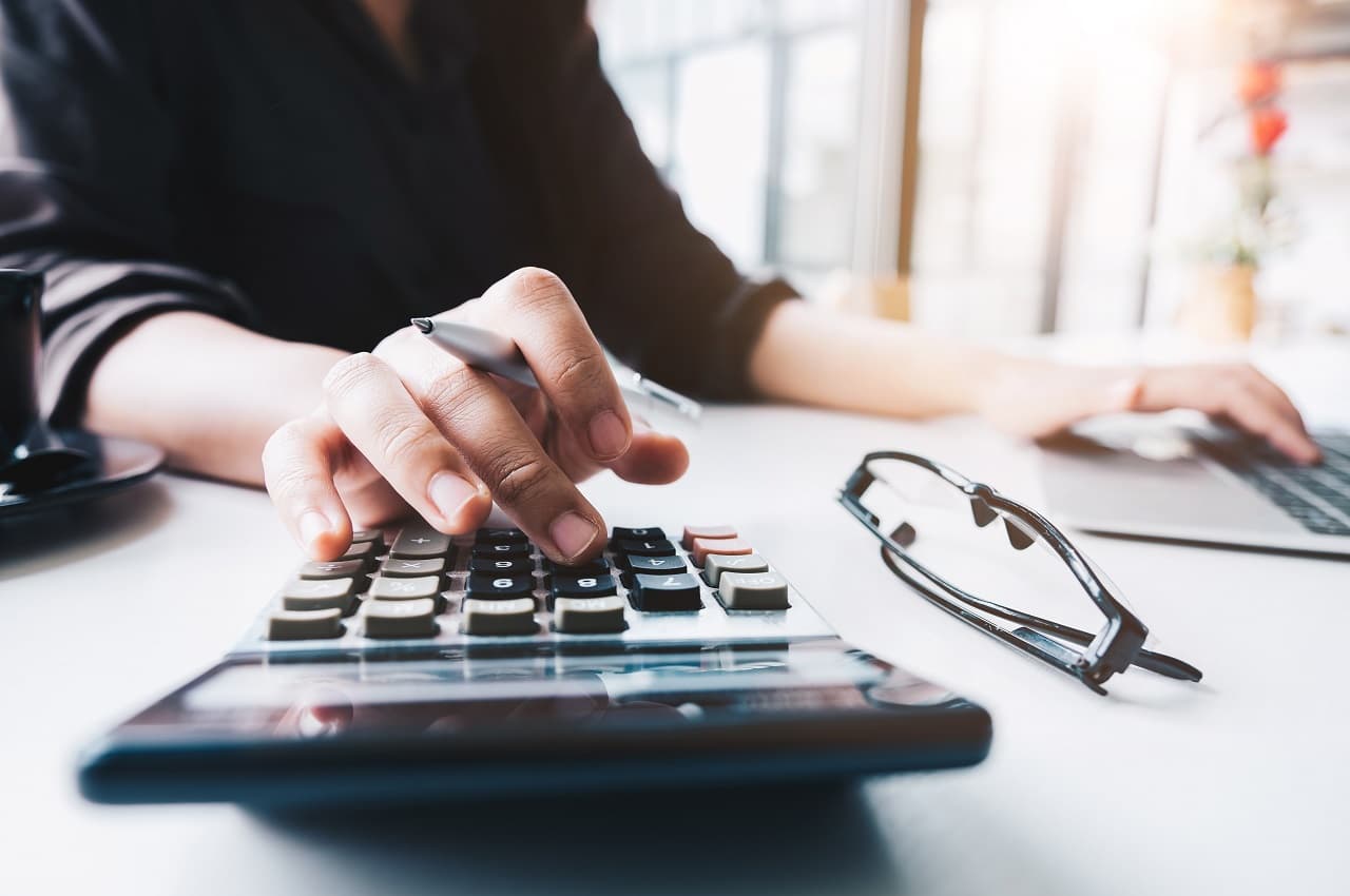 Foco em mãos de uma pessoa usando calculadora simples e, ao mesmo tempo, digitando em laptop em cima da mesa.