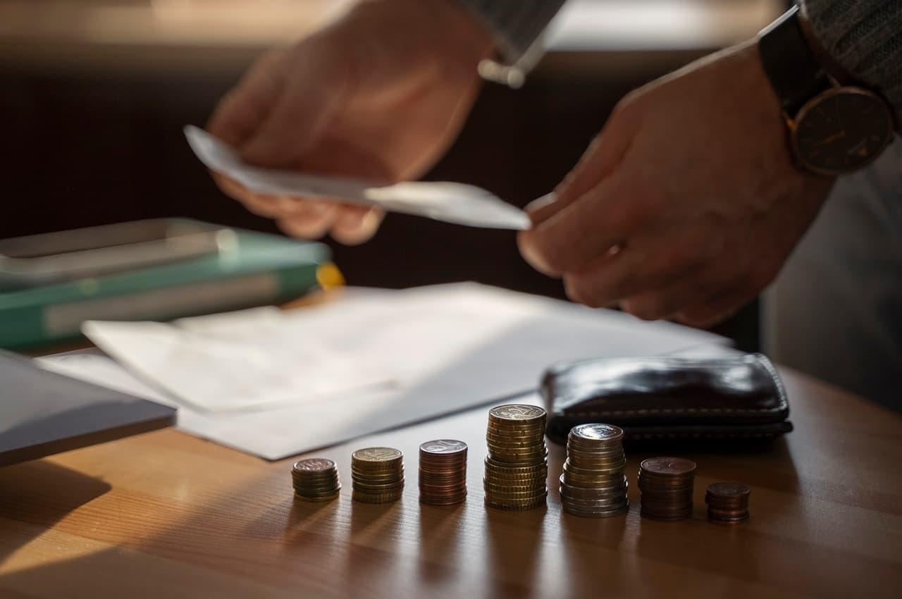 Pessoa (seu rosto não aparece na imagem) segura folha de papel de reajuste salarial, ao lado de uma pilha de moedas e um laptop aberto na mesa.