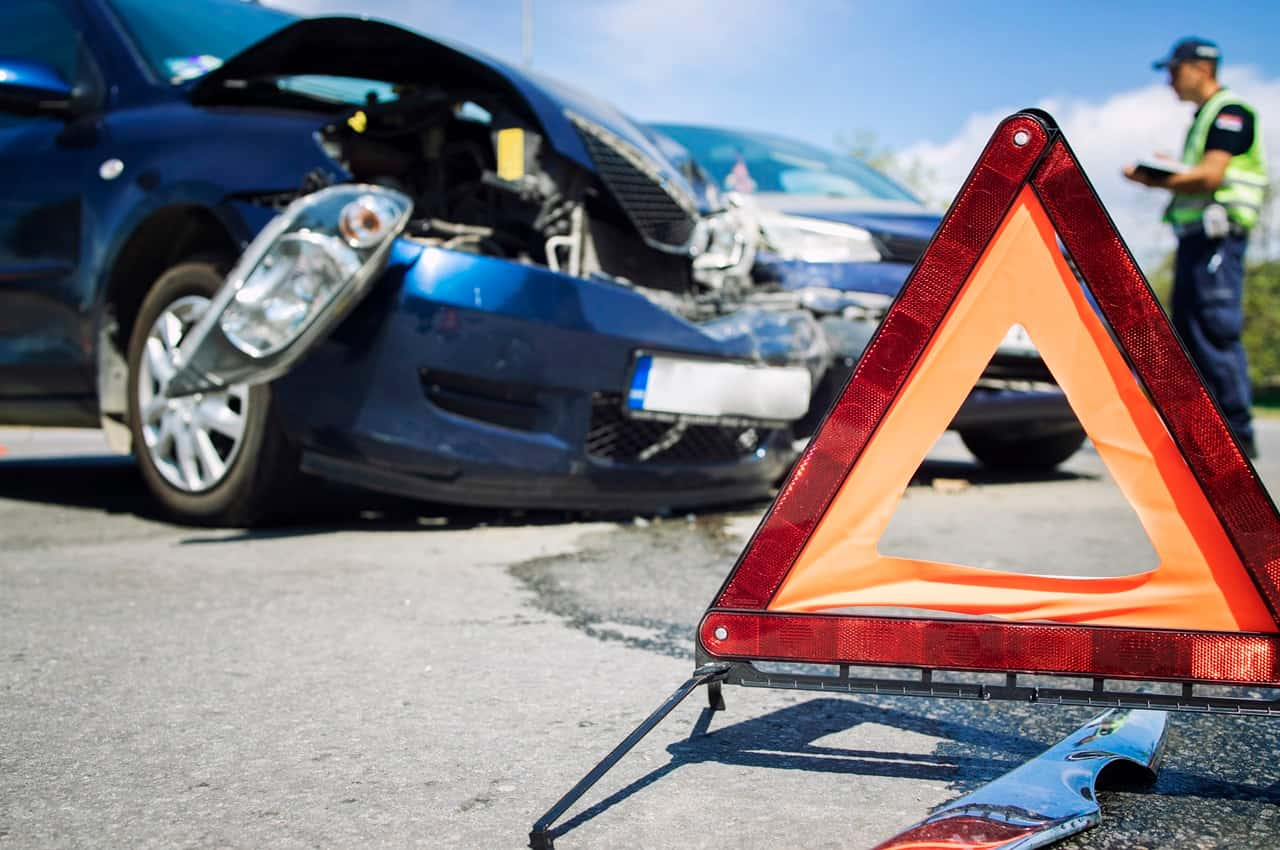 Veículo acidentado por uma batida de carro no modelo sedan e na cor azul escuro. Em frente ao veículo, temos um sinalizador triângulo e um guarda trânsito.