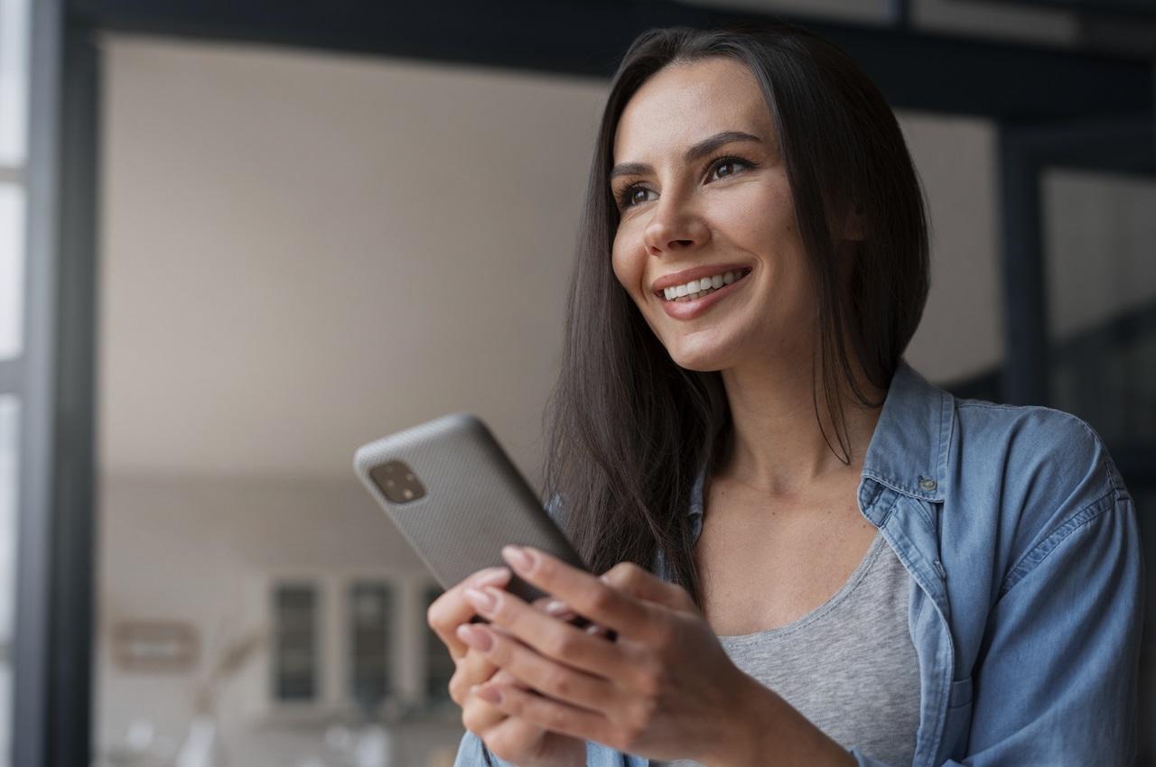 Mulher sorridente de cabelos longos castanhos e lisos, veste camiseta jeans e segura celular com as duas mãos para acessar empréstimo para CLT.