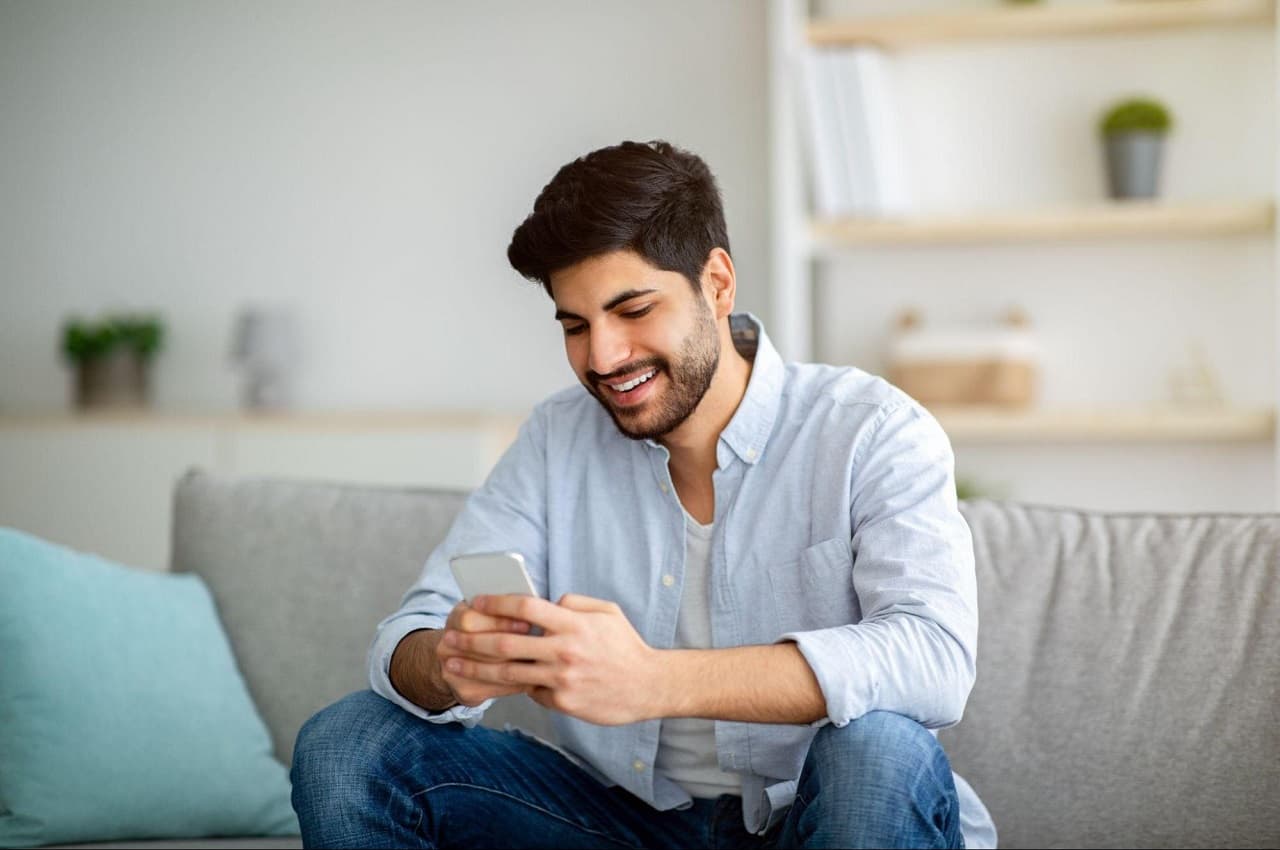 Homem sorridente sentado no sofá, digitando no celular e vestindo uma blusa social azul clara e calça jeans.